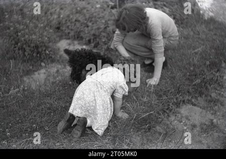 1950er Jahre, historisch, Mutter mit kleiner Tochter und Hund, kleines Mädchen, das auf dem Gras kniet und den Hund beobachtet, was Wasser hat, England, Großbritannien. Stockfoto