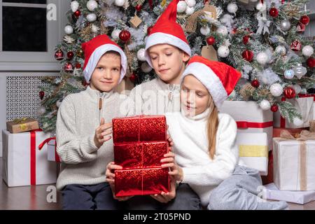 Drei süße, fröhliche, aufgeregte Kinder, Jungen und ein Mädchen mit weihnachtsmannmützen tauschen zu Hause Überraschungsgeschenke neben dem weihnachtsbaum aus. Schwester und Brüder geben Chri Stockfoto