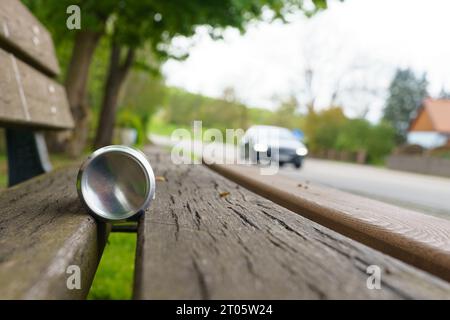 Auf der Bank steht eine leere Bierdose, und im unscharfen Hintergrund fährt ein Auto die Straße hinunter. Betrunkenes Fahrkonzept. Stockfoto