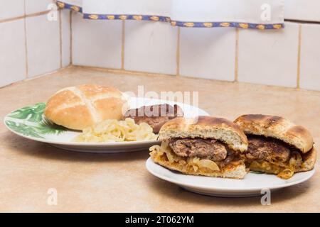 Ein zubereiteter Hamburger, der in zwei Hälften geschnitten wird, um sein saftiges hausgemachtes Patty zu enthüllen, auf einem Teller platziert wird, und ein anderer Teller zeigt einen unzubereiteten Hamburger, ungetoastet Stockfoto