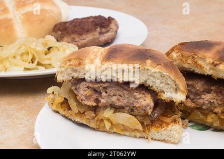 Eine Nahaufnahme eines hausgemachten Hamburger, der in zwei Hälften geschnitten ist und auf frisch gekochten Zwiebeln zwischen gerösteten Burgerbrötchen liegt. Im Hintergrund ist da Stockfoto