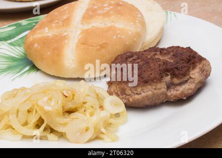 Eine köstliche Nahaufnahme mit geröstetem Hamburger-Patty, einem Burgerbrötchen und goldenen, durchscheinenden Zwiebeln auf einem weißen Teller. Burger-Moment. Stockfoto