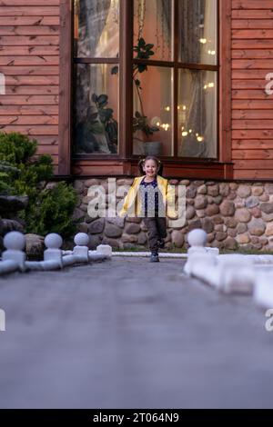 Wundervolle kleine Dame mit braunen Haaren in Pferdeschwänzen, in hellgelber Jacke, die auf der Straße zur Kamera läuft, mit fröhlichem Lächeln im Gesicht, s Stockfoto