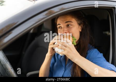 Die junge Frau sitzt im Auto und isst einen Burger. Fastfood im Auto Stockfoto