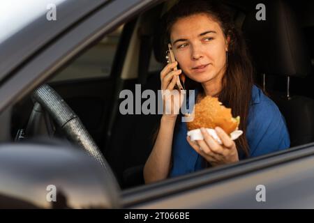 Frau isst einen Burger und spricht am Telefon, während sie im Auto sitzt. Fastfood und Take-away unterwegs. Stockfoto