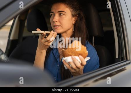 Nahaufnahme einer Frau, die ihr Handy benutzt, Burger isst und während des Autofahrens auf dem Lautsprecher spricht. Eine schöne Frau, die über das Freisprechtelefon im Auto spricht Stockfoto