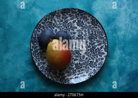 Ein Stillleben mit Birne und Pflaumen auf einem Teller mit Blumenmuster auf blauem Aquarellhintergrund Stockfoto