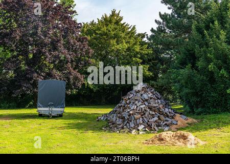 Ein großer Haufen gesägtem Brennholz und ein Autoanhänger auf einem grünen Rasen vor dem Hintergrund großer Bäume. Stockfoto