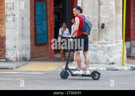 Ein junger Mensch mit Rucksack fährt einen Elektroroller in einer Stadtstraße. Andere Leute in schwarz-weißer Uniform stehen auf der Veranda eines Alten Stockfoto