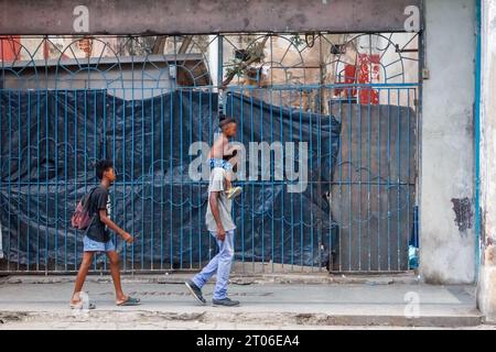 Ein kubanischer Mann trägt seinen Sohn in den Schultern, während er an einem eingestürzten Gebäude vorbeigeht. Die Ruine wurde durch eine Metallgittertür geschützt Stockfoto