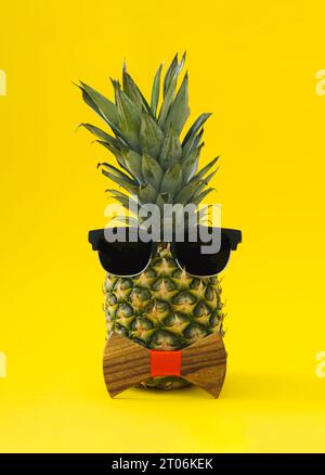 Kreatives tropisches Sommerlayout aus Ananas, Sonnenbrille und hölzerner Fliege auf hellgelbem Hintergrund. Minimales Sommerfrucht-Konzept. Stockfoto
