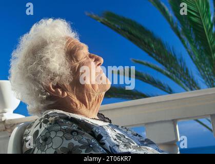 Ältere Frau im Urlaub entspannt und sonnt sich zufrieden in der warmen Sonne in ihrem Ferienhaus, auf ihrer Terrasse mit Palme hinter den Kanarischen Inseln Spanien Stockfoto