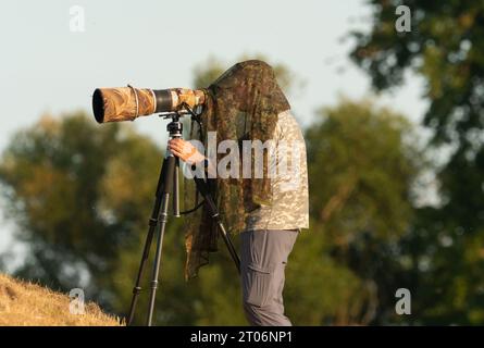 Lutherstadt Wittenberg, Deutschland. September 2023. 28.09.2023. Ein Naturfotograf steht auf einem Deich bei den Elbwiesen an der Elbe, etwa zehn Kilometer südöstlich von Lutherstadt Wittenberg, und blickt durch seine Kamera, an der ein leistungsstarkes Teleobjektiv befestigt ist. Über seinem Kopf trägt der Mann ein Tarnnetz, denn wilde Tiere und seltene Vögel erkennen Menschen besonders gut an ihrer Silhouette und ihrem Gesicht. Quelle: Wolfram Steinberg/dpa/Alamy Live News Stockfoto