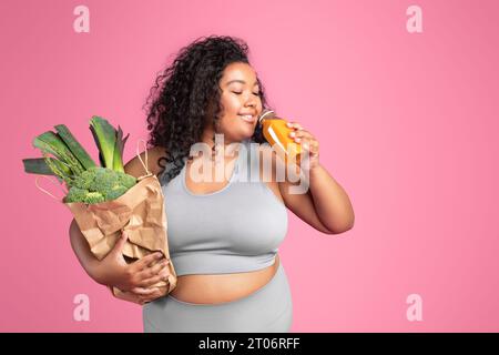 Positive schwarze mollige Frau in Sportbekleidung, die einen Beutel mit grünem Gemüse hält und frischen Smoothie trinkt, freier Platz Stockfoto