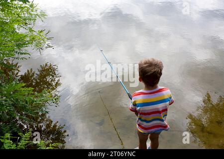 Ein Vorschulkind fischt am bewölkten Sommertag im Fluss. Junge warf Angelrute, steht mit dem Rücken zur Kamera, Wolken spiegeln sich im Fluss. Natur Stockfoto