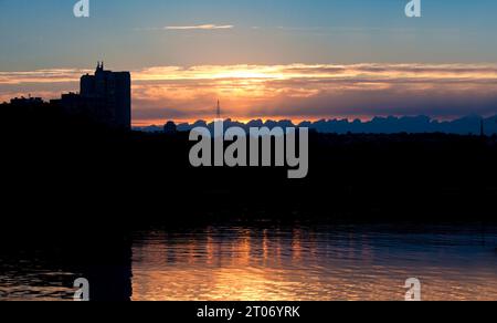 Urbane Landschaft. Sonnenuntergang in der Stadt. Silhouetten von Häusern vor dem Hintergrund der untergehenden Sonne und des untergehenden Himmels spiegeln sich im dunklen Fluss wider. Ukraine, Dnipro Stockfoto