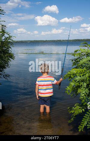 Ein Vorschulkind fischt am sonnigen Sommertag im Fluss. Der Junge wirft eine Angelrute, steht im Wasser mit dem Rücken zur Kamera, ein Blick auf den Fluss, Wolken hinein Stockfoto