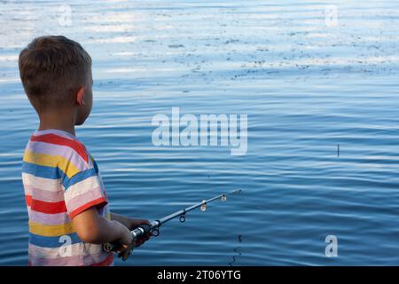 Ein Vorschulkind fischt am Sommertag im Fluss. Junge hat Köder geworfen und wartet auf Biss, stehend mit dem Rücken zur Kamera. Amateurfischen in o Stockfoto