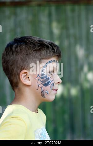 Profilporträt eines Vorschuljungen mit gemaltem Skorpion im Gesicht. Festliche Gesichtsmalerei. Glückliches braunäugiges Kind draußen auf grünem Hintergrund. Summe Stockfoto