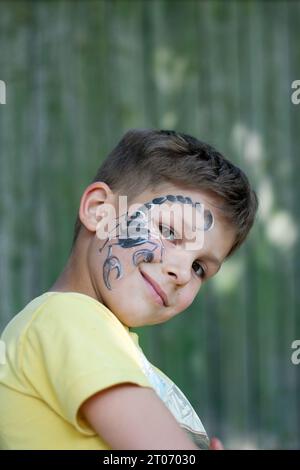 Porträt eines Vorschuljungen mit gemaltem Skorpion im Gesicht. Festliche Gesichtsmalerei. Glückliches braunäugiges Kind draußen auf grünem Hintergrund. Sommer. Körper Stockfoto