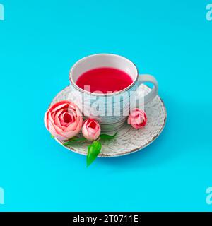 Kreatives Layout aus einer Tasse Tee, dekoriert mit rosa Blumen und grünen Blättern auf hellblauem Hintergrund. Minimales Heißgetränk- und Blumenkonzept Stockfoto