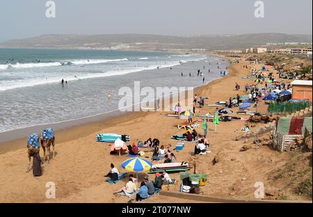 Surfer, Kamele und Sonnenanbeter genießen einen Februartag am Strand von Tamraght in Taghazout Bay bei Agadir in Marokko. Stockfoto