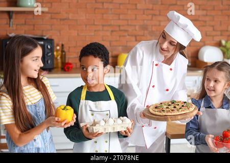 Weiblicher Koch mit zubereiteter Pizza und eine Gruppe kleiner Kinder nach dem Kochkurs in der Küche Stockfoto