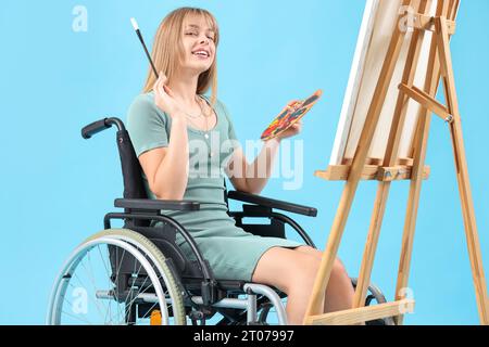 Junge Frau im Rollstuhlmalerei auf Staffelei vor blauem Hintergrund Stockfoto