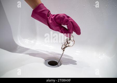 Hand der Frau in rosafarbenem Gummihandschuh, der Haarklumpen vom Wasserablauf abnimmt, verstopfte Leitung im Bad. Stockfoto