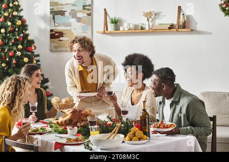 Fröhliche multirassische Familie, die viel Spaß beim festlichen Mittagessen hat, Wein trinkt und truthahn essen, Weihnachten Stockfoto