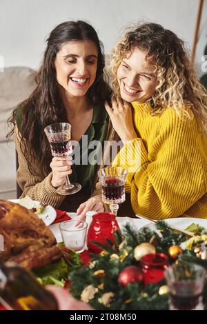 Fröhliches lgbt-Paar, das sich umarmt und aufrichtig lächelt, Weingläser hält, Weihnachten Stockfoto