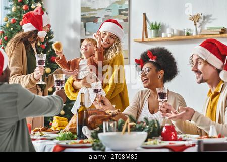 Große multikulturelle und fröhliche Familie in Weihnachtsmützen, die ihre Weingläser am Weihnachtstisch anstoßen Stockfoto