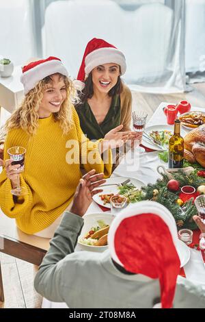 Multikulturelle Familie in lässiger Kleidung und Weihnachtsmannmützen sitzen am Weihnachtstisch und unterhalten sich lebhaft Stockfoto