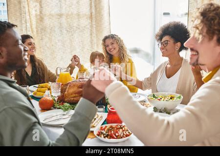 Fröhliche multirassische Freunde und Familie halten Hände und beten zusammen am Thanksgiving-Tisch Stockfoto