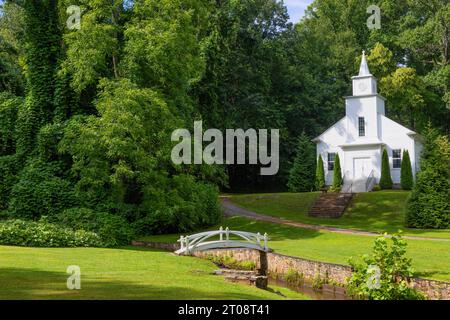 Lure Lake, North Carolina, USA - 11. August 2023: Schöne weiße Kirche mit einer Straße und einem Bach mit Fußgängerbrücke im Vordergrund. Stockfoto