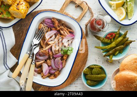 Gezapftes bbq-Hühnchen mit gebackenen Zwiebeln auf Servierplatte, bbq-Mais, Gurken, Chilischoten und Brötchen für Hot Dogs und Burger, Tomatensauce. Traditionell Stockfoto