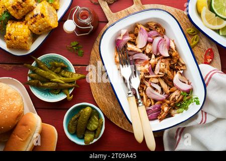 Gezapftes bbq-Hühnchen mit gebackenen Zwiebeln auf Servierplatte, bbq-Mais, Gurken, Chilischoten und Brötchen für Hot Dogs und Burger, Tomatensauce. Traditionell Stockfoto