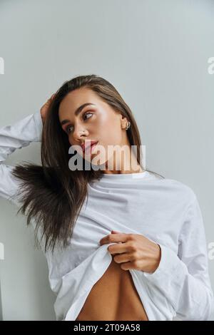 Schlanke Frau mit langärmligen Hemden und langem Haar, die sich in die Nähe der weißen Wand schaut Stockfoto