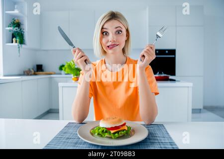 Foto von verwirrten hübschen Frauenhänden halten Gabelmesser beißen Lippen Burgerteller Küchentisch helles Haus innen Stockfoto