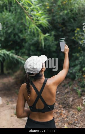 Anonyme Frau mit Sportswear lächelt und nimmt Selfie per Smartphone, während sie im Wald steht Stockfoto