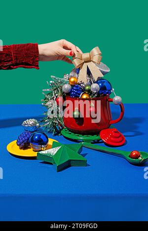 Beschneiden Sie anonymes Weibchen mit roten Nägeln, die weihnachtsbandkegel in einen Topf mit verschiedenen Ornamenten neben Teller und Löffel gegen blaues und grünes Backgro legen Stockfoto