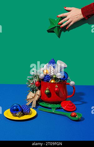 Ernten Sie anonymes Weibchen mit roten Nägeln, die einen grünen weihnachtsstern über dem Topf halten, mit verschiedenen Ornamenten in der Nähe des Tellers und des Löffels gegen blauen und grünen Hintern Stockfoto