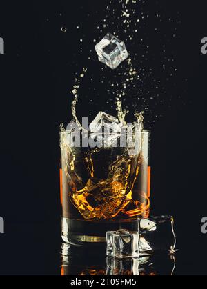 Ein Glas Whisky, Cognac oder Rum mit 3 Eiswürfeln auf schwarzem Hintergrund macht einen großen Spritzer mit vielen Tropfen Stockfoto