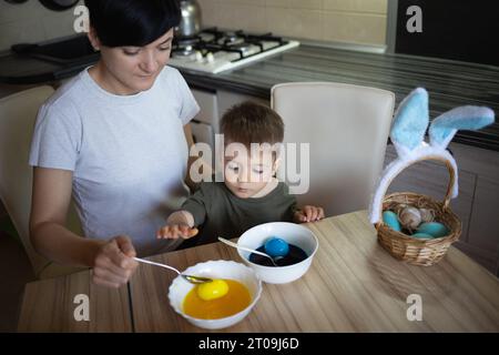 Kleiner süßer Junge mit Mutter malt Eier für ostern, die in der Küche am Tisch sitzen Stockfoto