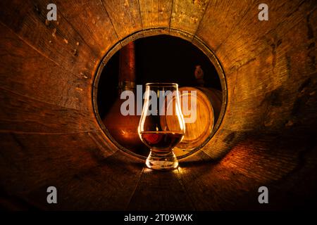 Ein Glas Scotch Whiskey in altem Eichenfass. Kupfer-alambic und kleines Fass auf Hintergrund. Traditionelles Alkoholdestilleriekonzept Stockfoto