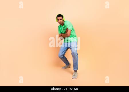 Foto in voller Größe von stilvollem Guy gekleidet grün T-Shirt Denim Hosen posiert in Kopfhörern Arme überkreuzt isoliert auf beigefarbenem Hintergrund Stockfoto