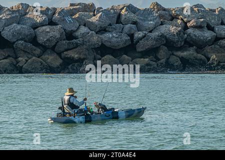 Ventura, CA, USA - 14. September 2023: Nahaufnahme. Einsamer Fischer in einer kleinen Schaluppe, die auf dem grünlichen Wasserweg am Eingang zum Hafen beschäftigt ist. Riesiger Pier aus grauen Felsen Stockfoto