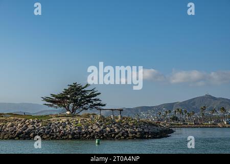 Ventura, CA, USA - 14. September 2023: Soter's Point Marina Park Baum und Bänke über Felsen bilden Pier unter blauem Himmel. Bergkette und Innenstadt Stockfoto