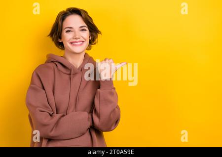 Foto des lustigen netten Mädchens zeigen Daumen Finger leeren Raum Billboard Mockup Kleidung braune Kapuze Hemd isoliert gelbe Farbe Hintergrund Stockfoto