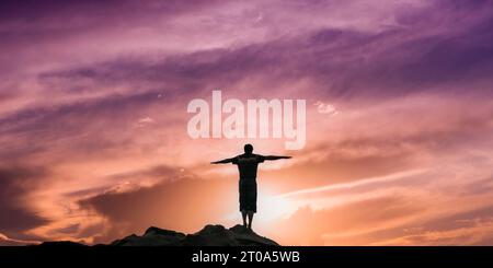 Ein Mann steht mit ausgestreckten Armen auf einem Berggipfel, seine Silhouette vor einem faszinierenden Sonnenuntergang in Flieder- und Sandtönen. Es ist ein Stockfoto